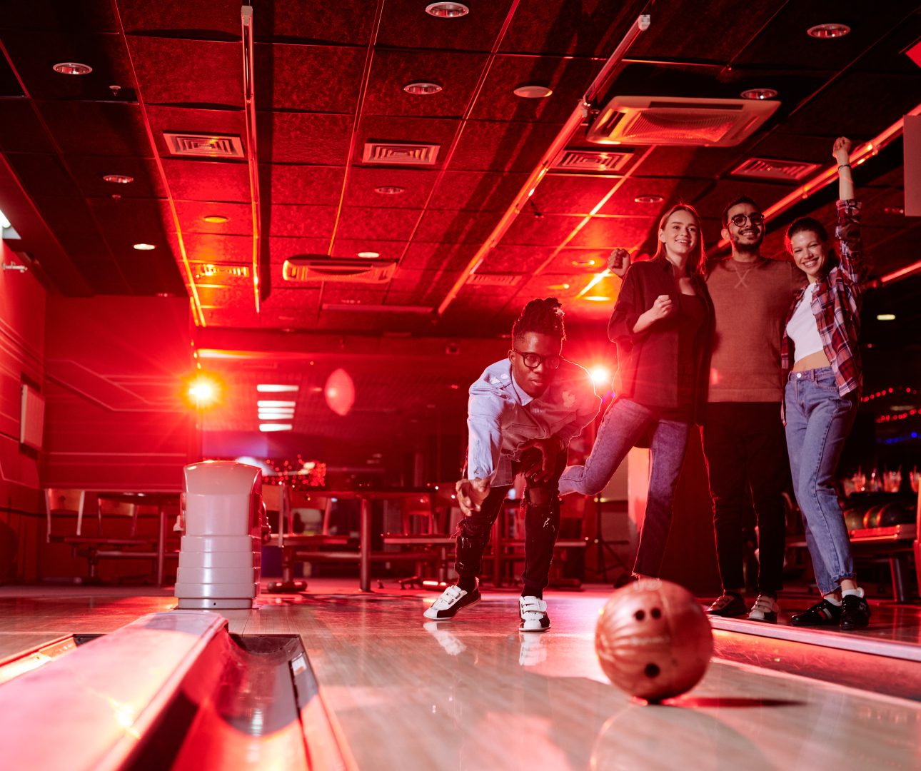 African guy bending by bowling alley while throwing ball on background of his friends with raised arms expressing gladness